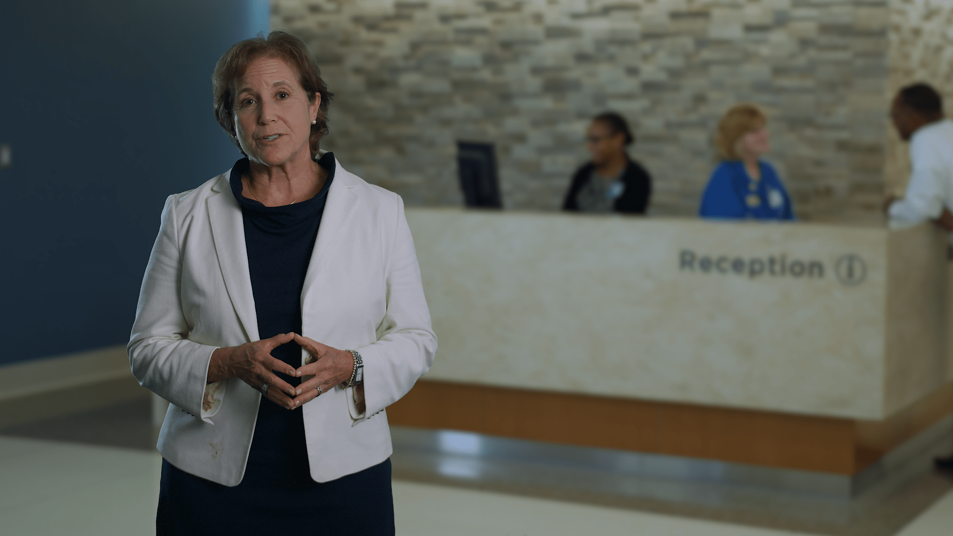 A still of a woman speaking in front of a reception desk. It's an example of a background plate. 